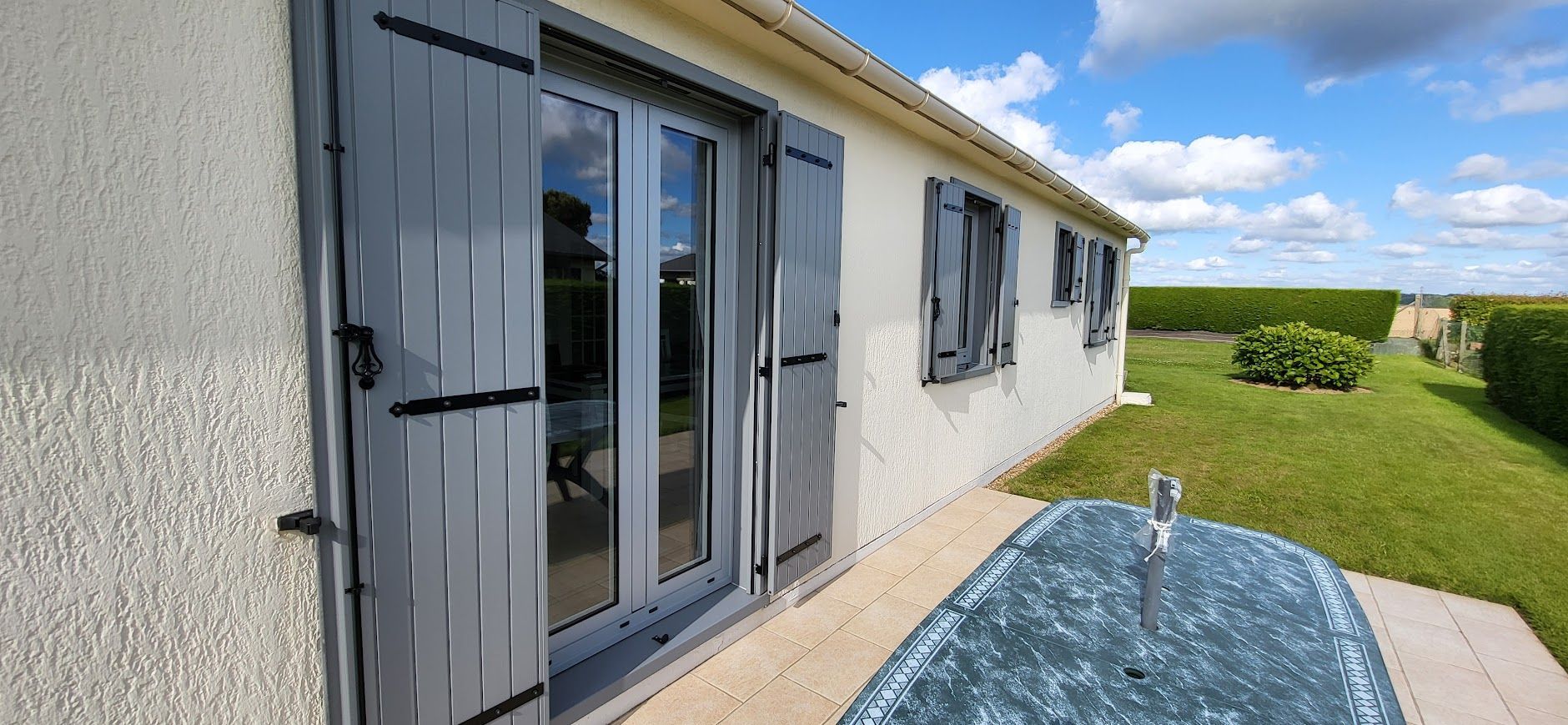Une maison aux volets gris et aux portes vitrées, située sur une pelouse avec une table.