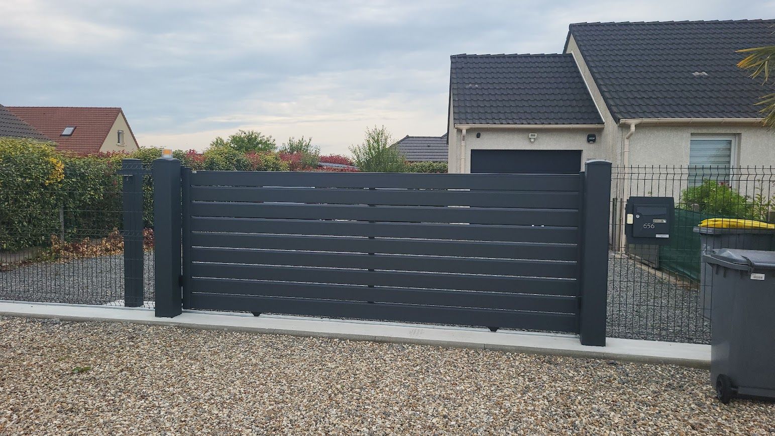 Clôture à lattes horizontales grises avec poteaux foncés, allée de gravier et maisons en arrière-plan.