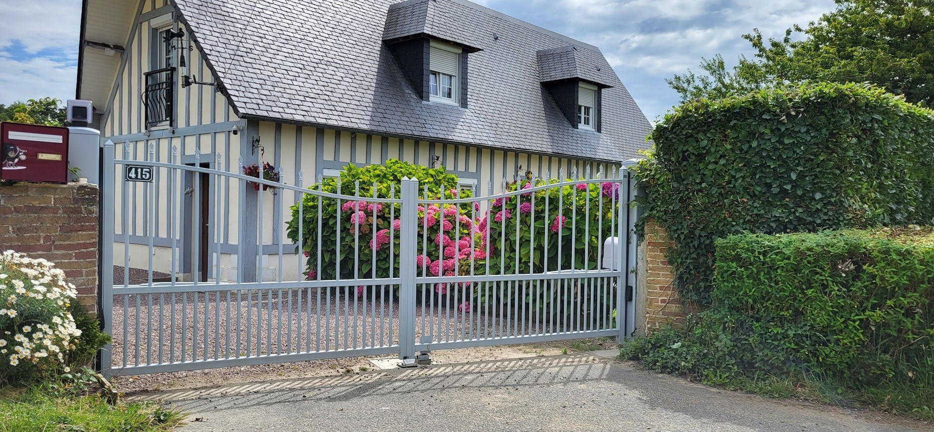 Une maison claire au toit sombre se cache derrière une clôture métallique grise. Des buissons verts et du gravier l'entourent.