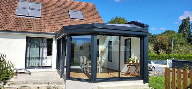 Une véranda moderne avec de grandes fenêtres est attenante à la maison. Ciel bleu au-dessus.