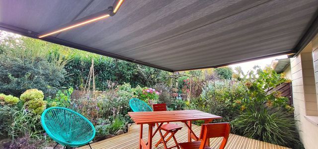 Une terrasse extérieure ombragée avec une terrasse en bois, une table et des chaises. Un jardin verdoyant luxuriant se dessine à l'arrière-plan.