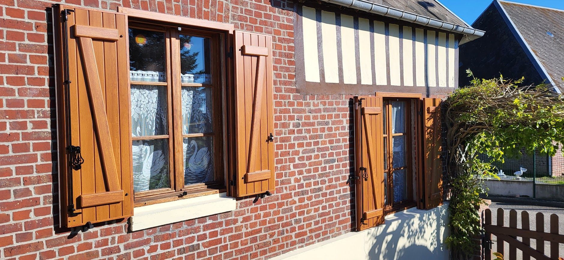 Une maison en briques avec des volets bruns à deux fenêtres, des vignes poussant près de la deuxième fenêtre.