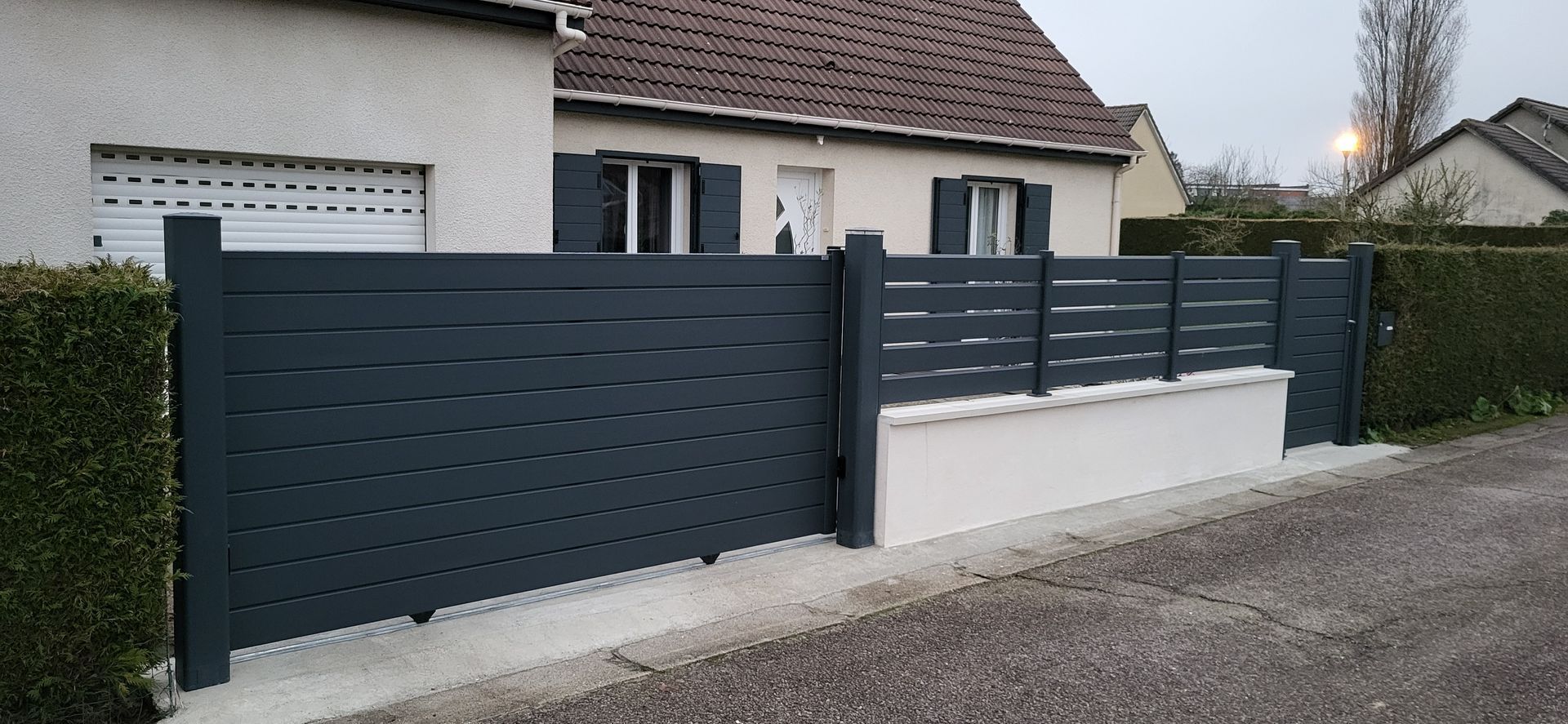 Clôture à lattes horizontales grises devant une maison. Pansement de mur blanc, haie verte, route grise.