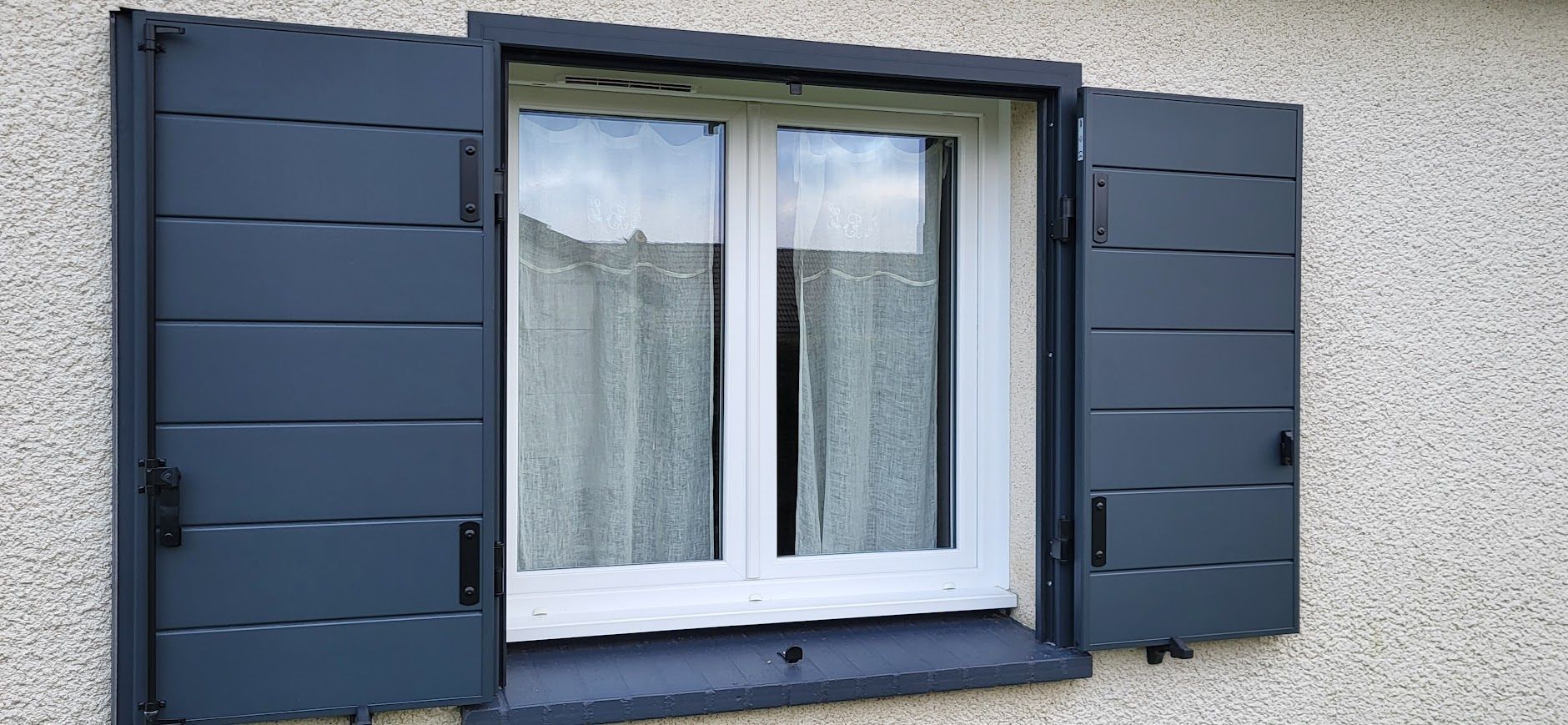 Fenêtre à volets gris foncé, ouverte des deux côtés. Une fenêtre à cadre blanc est visible derrière des voilages.