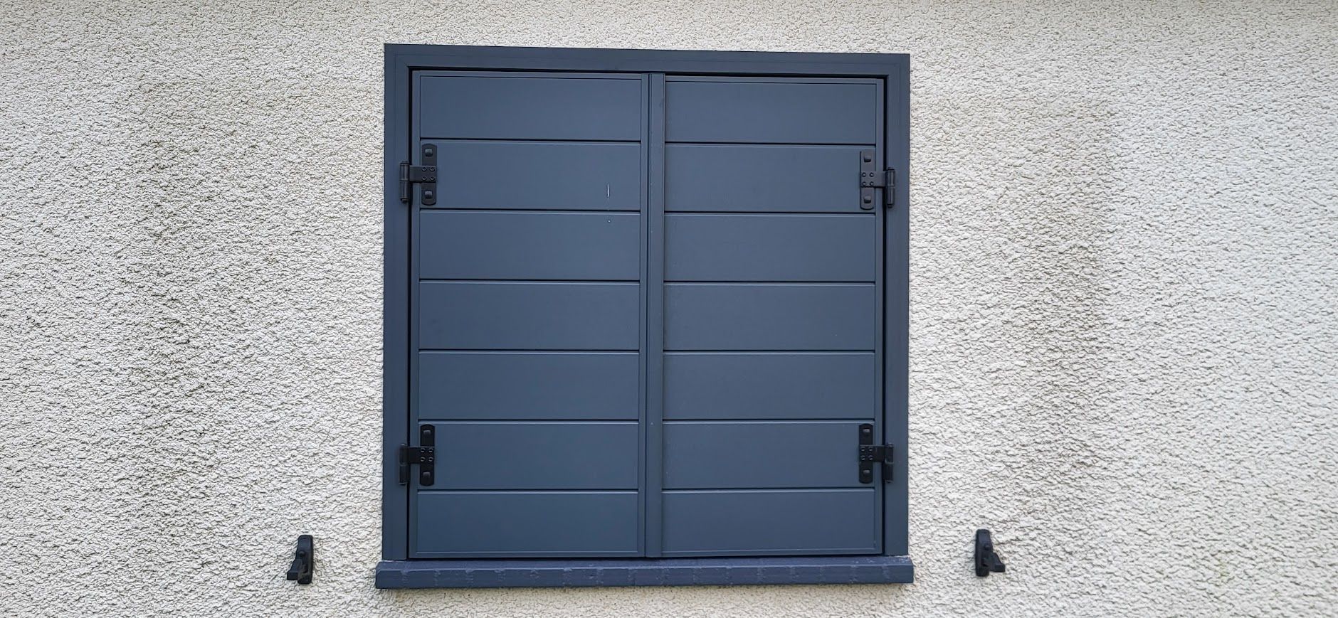 Volets gris sur un mur en stuc. Charnières et supports noirs.