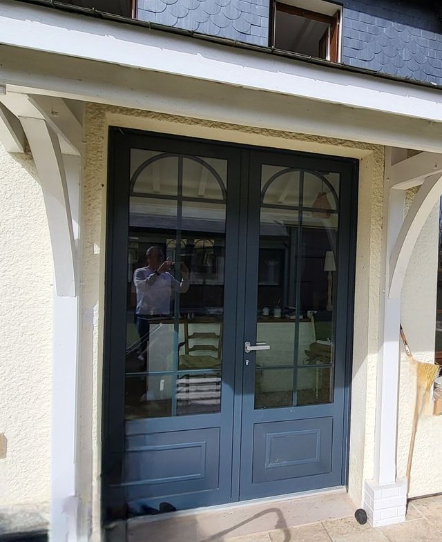 Double porte bleue à panneaux de verre cintrée, sous un porche blanc. Une personne se reflète dans le verre.