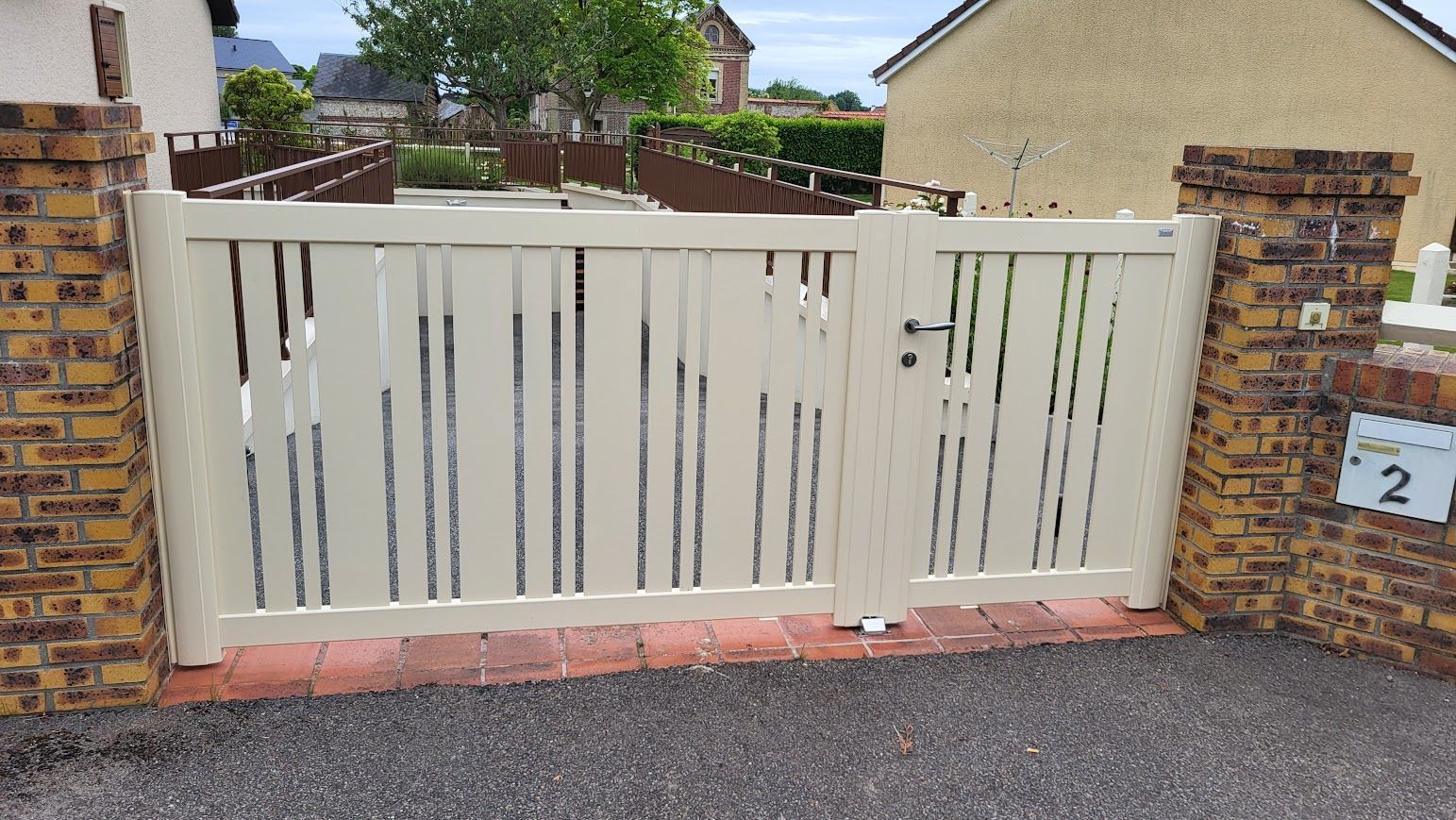 Portail en métal beige avec découpes verticales entre des piliers en briques, donnant sur une allée.