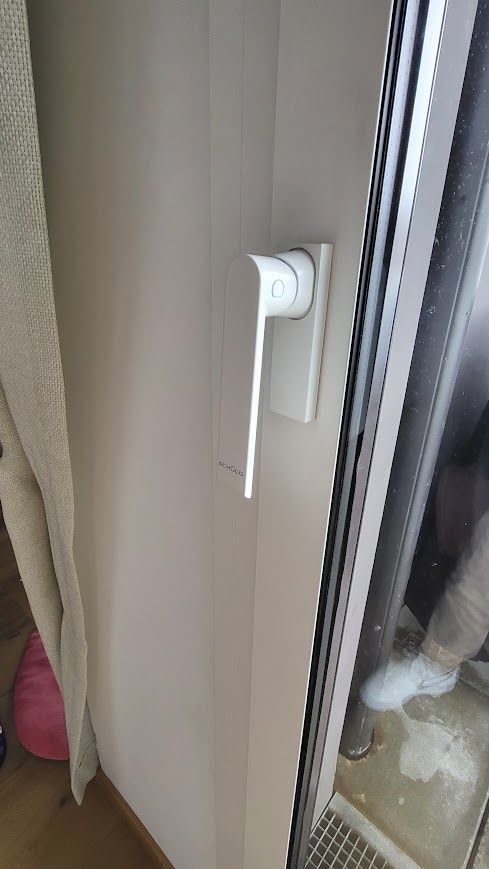 Poignée de fenêtre blanche sur un mur beige, à côté d'une fenêtre.