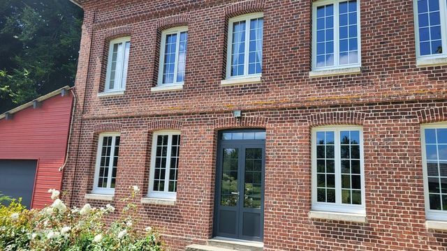 Maison en briques aux fenêtres à cadre blanc et à porte sombre, située dans un paysage verdoyant.