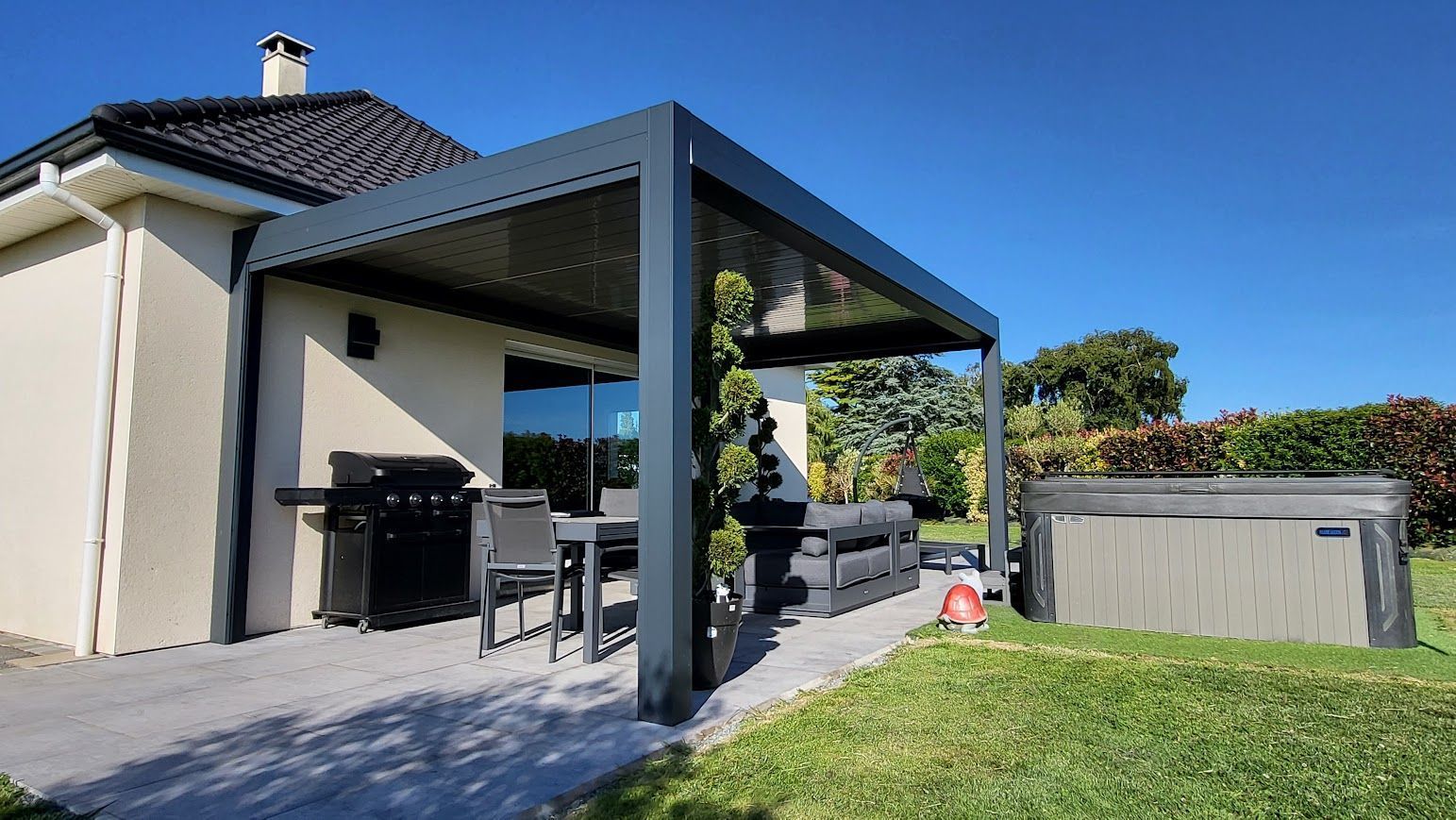 Terrasse avec pergola, barbecue, table, jacuzzi et maison se détachant sur un ciel bleu azur.