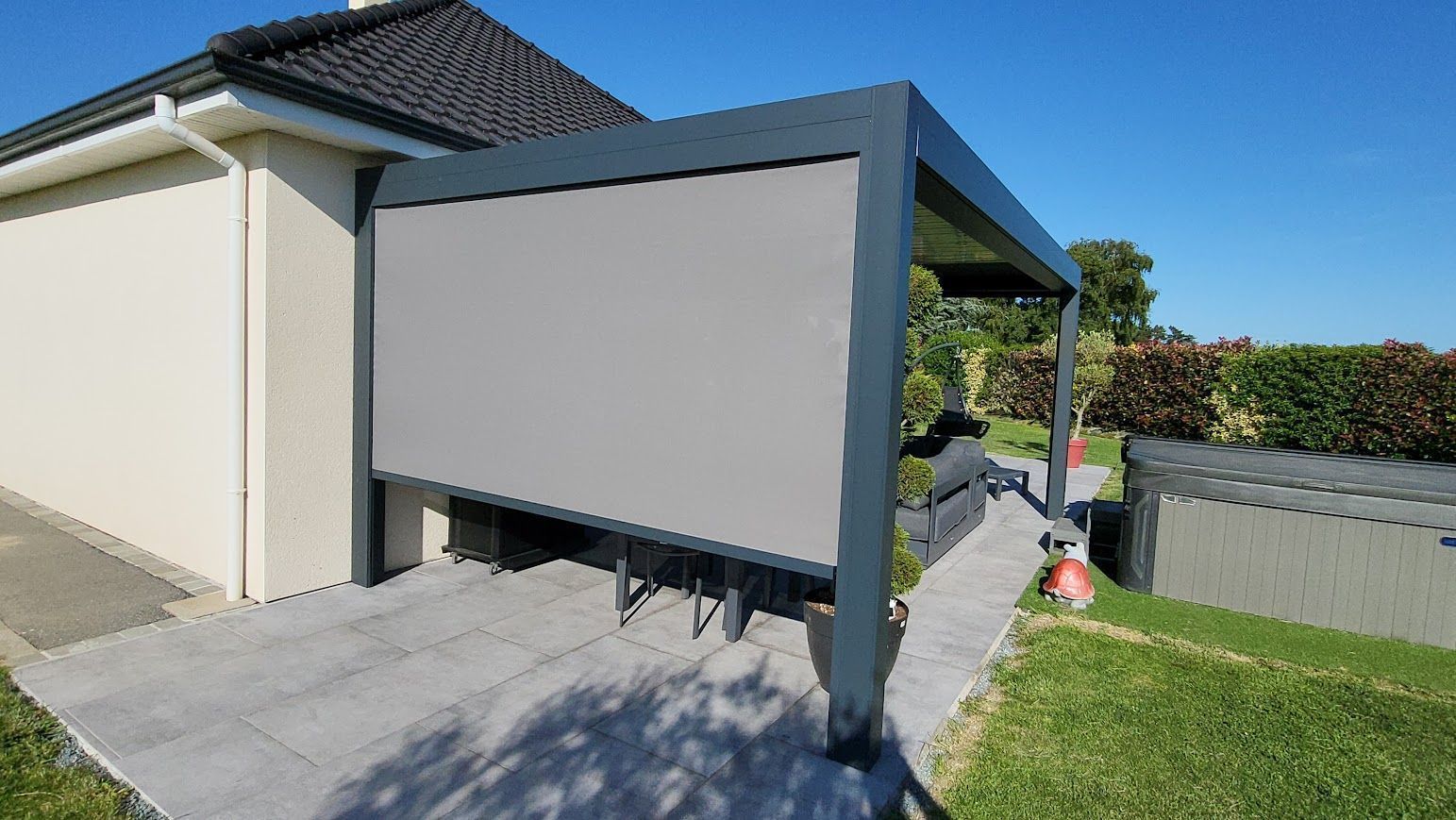 Un auvent gris, fixé à une maison et muni d'un écran abaissé, offre de l'ombre sur une terrasse.