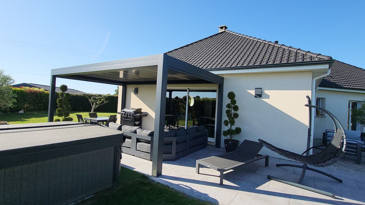 Patio avec pergola grise, mobilier de jardin et maison sur fond de ciel bleu.