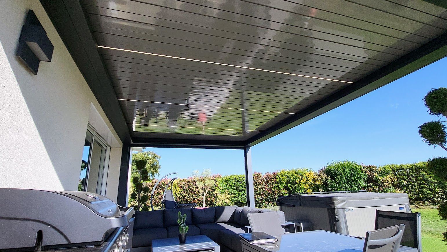 Patio avec toit en pergola grise, mobilier de jardin et vue sur la verdure et le ciel bleu.