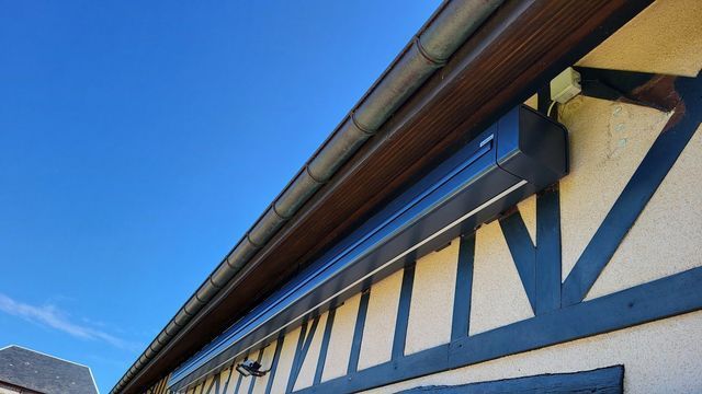 Un auvent noir est installé sur une maison aux boiseries brunes, sous un ciel bleu.