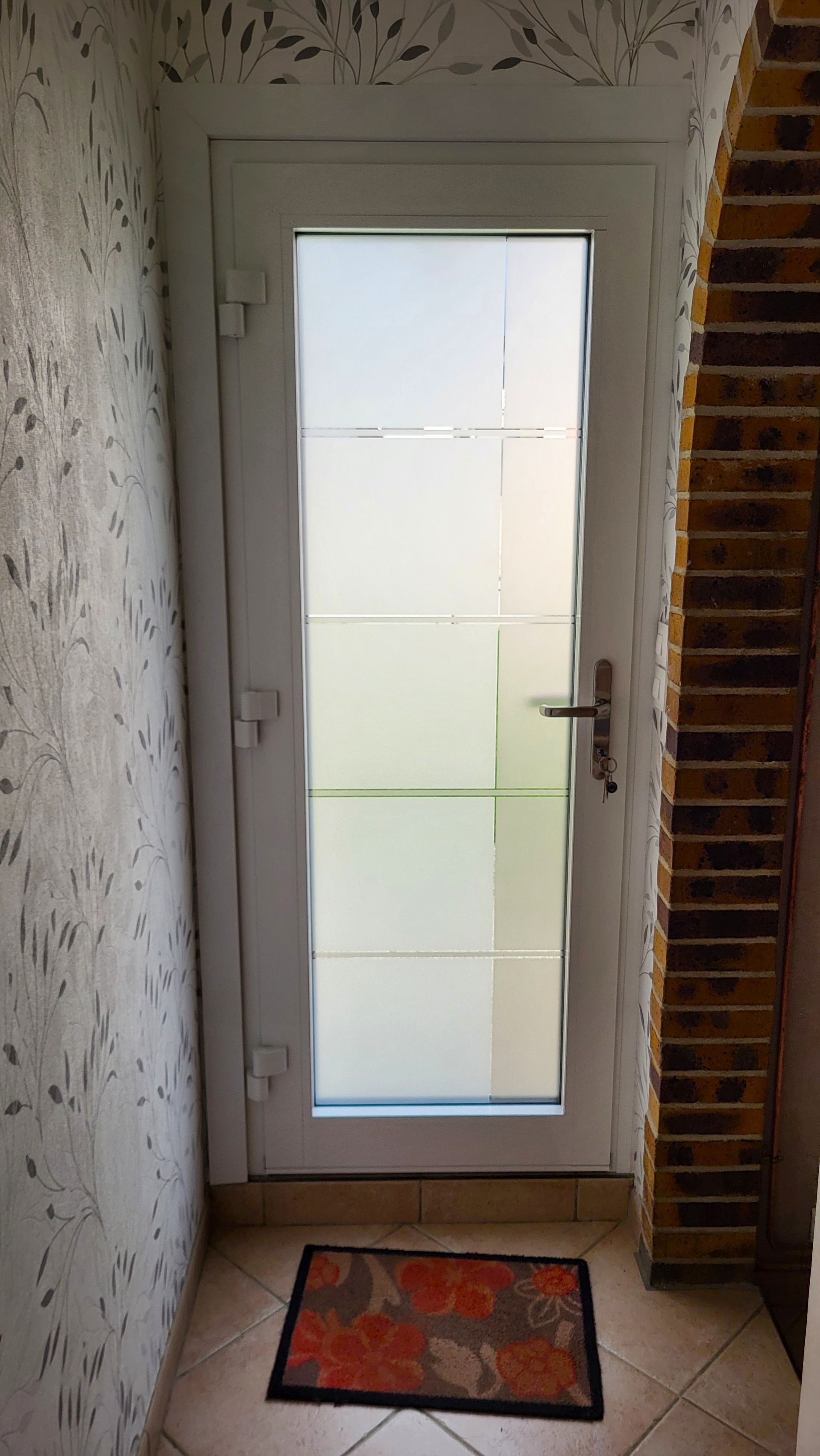 Porte blanche avec panneaux de verre dépoli, paillasson sur sol carrelé, arche en briques à droite.
