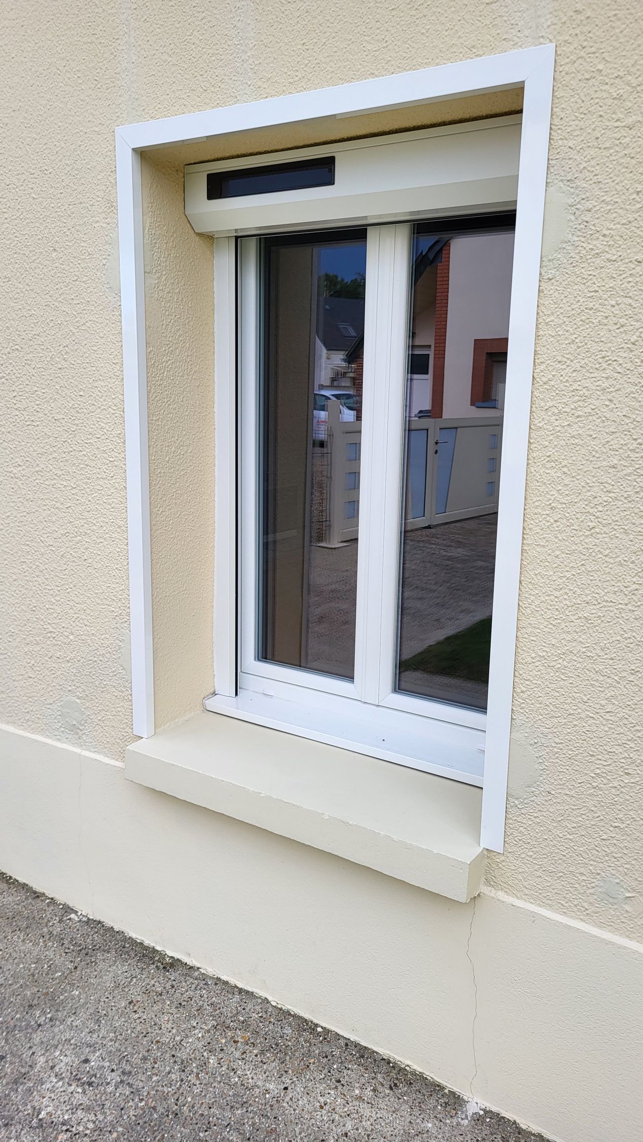 Fenêtre à cadre blanc, stores fermés, sur façade en stuc beige.