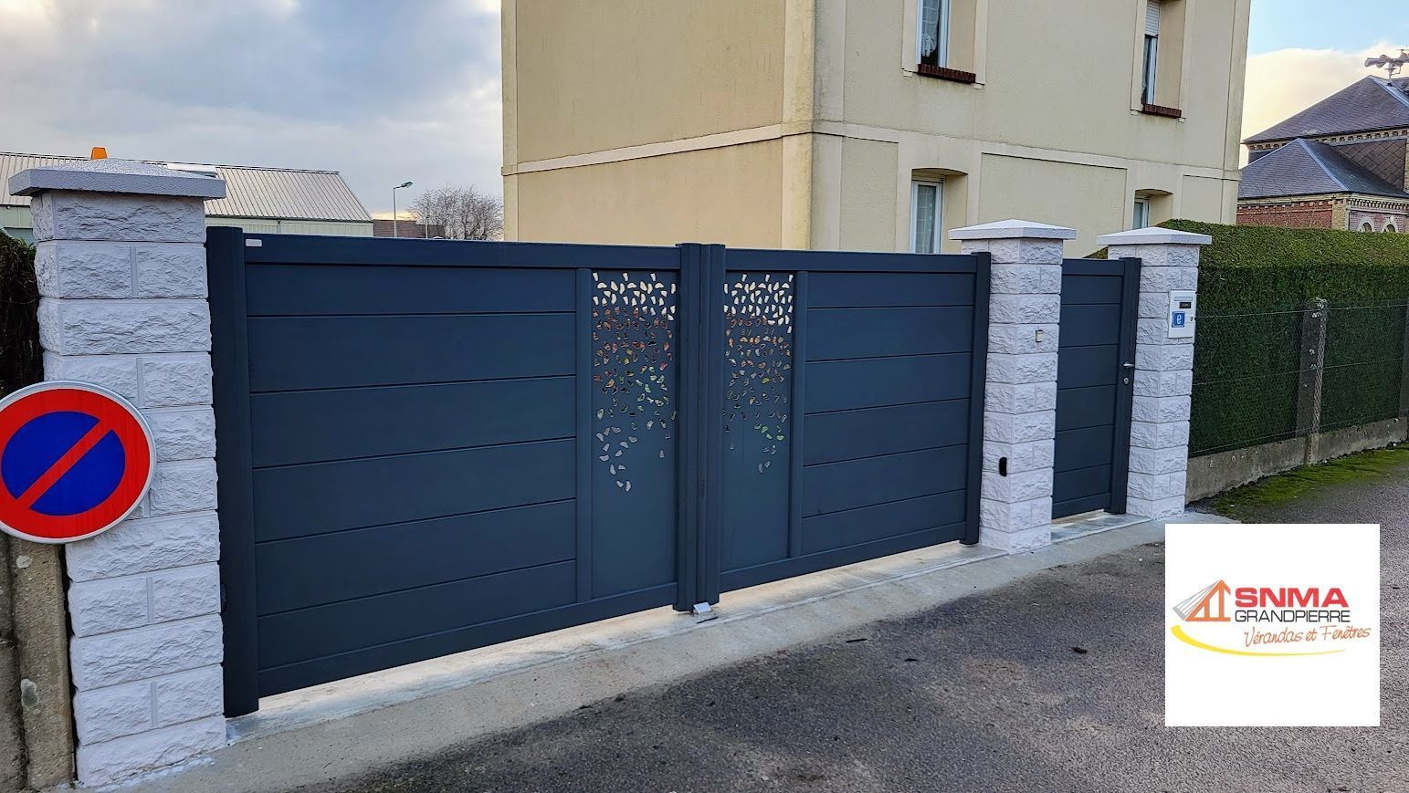 Portail d'allée en métal gris foncé avec découpes décoratives, placé entre des piliers en pierre grise.