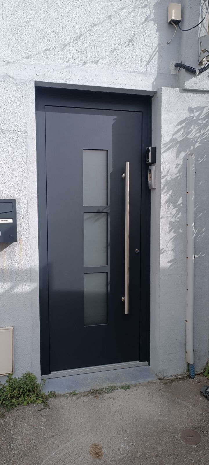 Une porte d'entrée gris foncé à trois panneaux de verre et dotée d'une longue poignée verticale, sur un mur texturé.