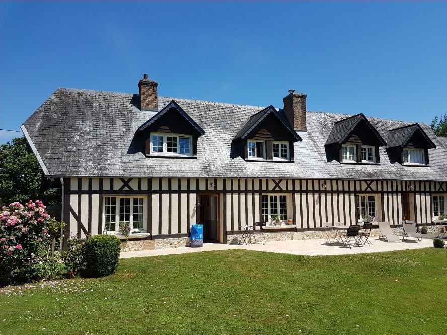 Maison à colombages avec lucarnes, boiseries blanches et toit d'ardoise, située sur une pelouse verte se détachant sur un ciel bleu.