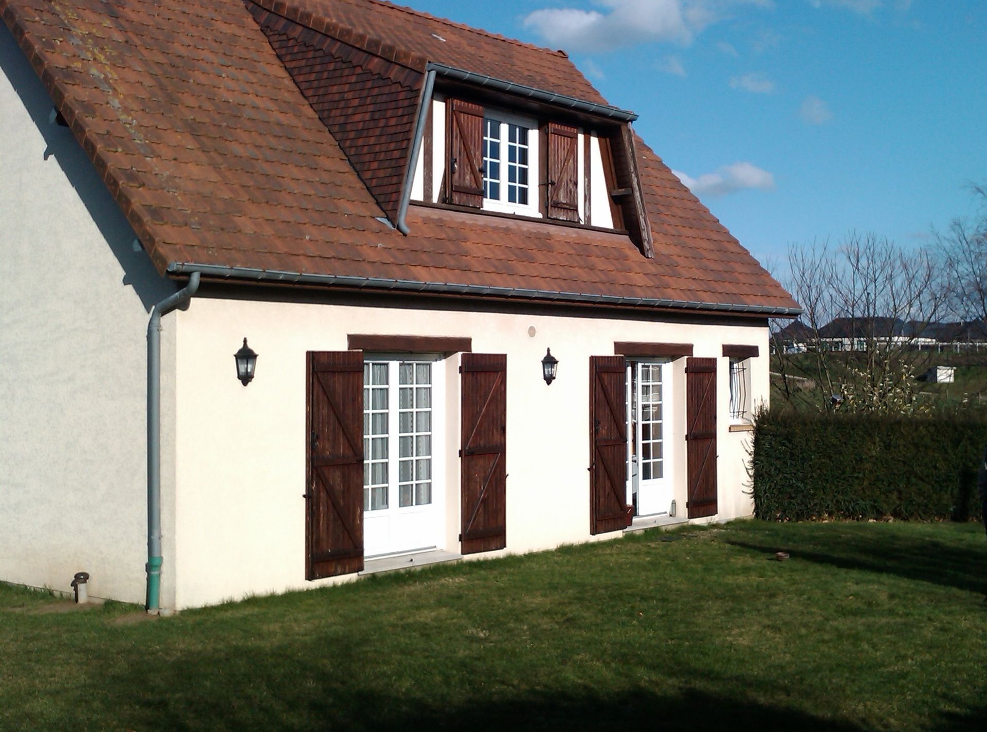 Un cottage au toit marron, aux murs blancs, aux volets marron et à la pelouse verte sous un ciel bleu.