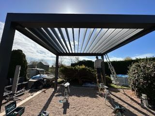 Pergola noire en construction, journée ensoleillée. Deux personnes, outils, sol de gravier et verdure.