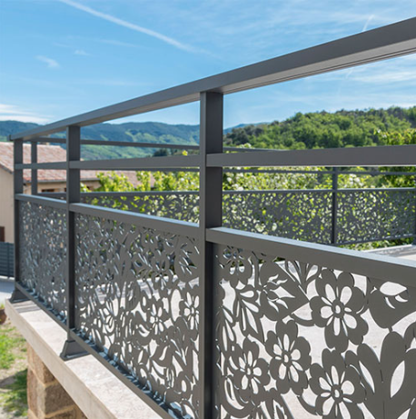 Rambarde de balcon en métal gris foncé ornée de découpes florales, donnant sur un paysage verdoyant par une journée ensoleillée.
