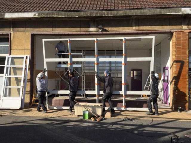 Des ouvriers installent un grand cadre de fenêtre blanc dans une ouverture du bâtiment.