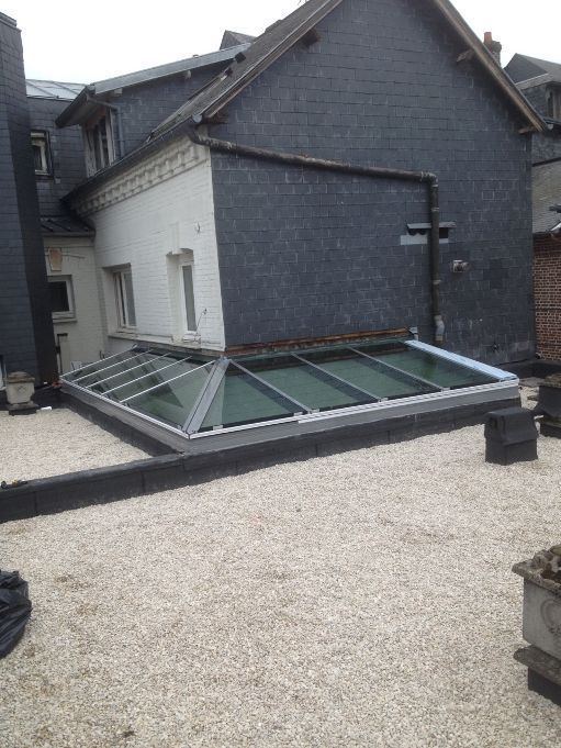 Puits de lumière sur le toit d'un immeuble aux murs blancs et gris foncé, entouré de petites pierres blanches.