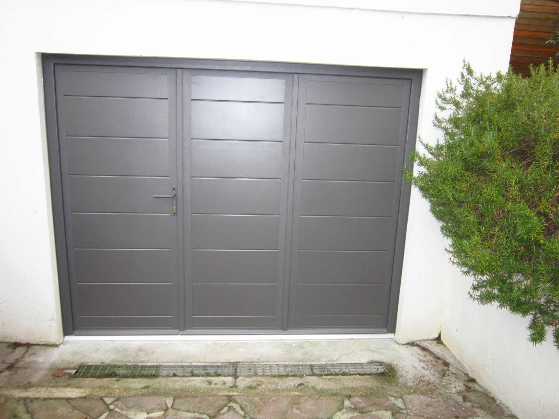 Porte de garage grise à panneaux horizontaux encastrée dans un mur blanc, à côté d'un buisson vert.