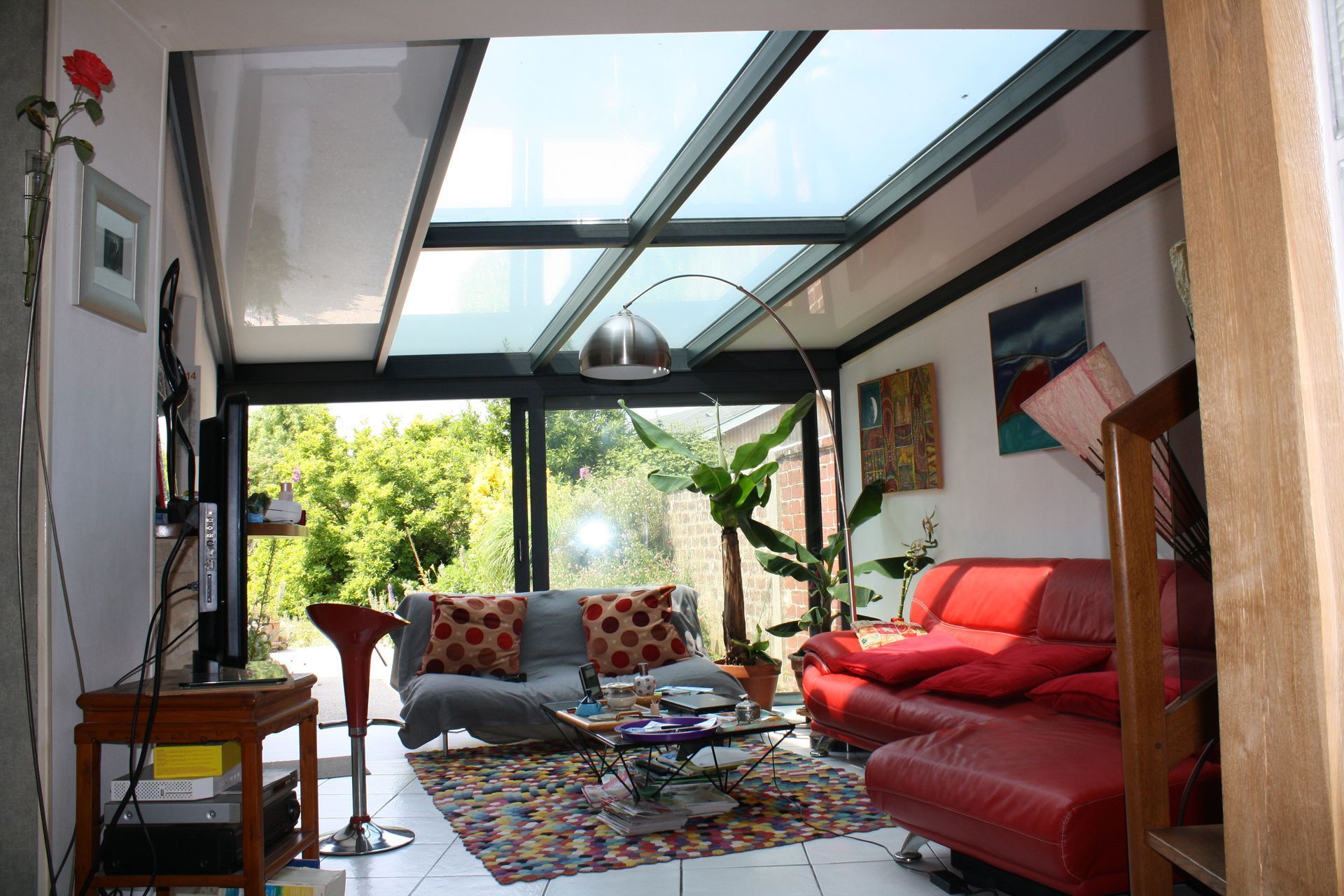 Véranda avec toit en verre, canapés rouges et gris, tapis coloré, vue sur la verdure.