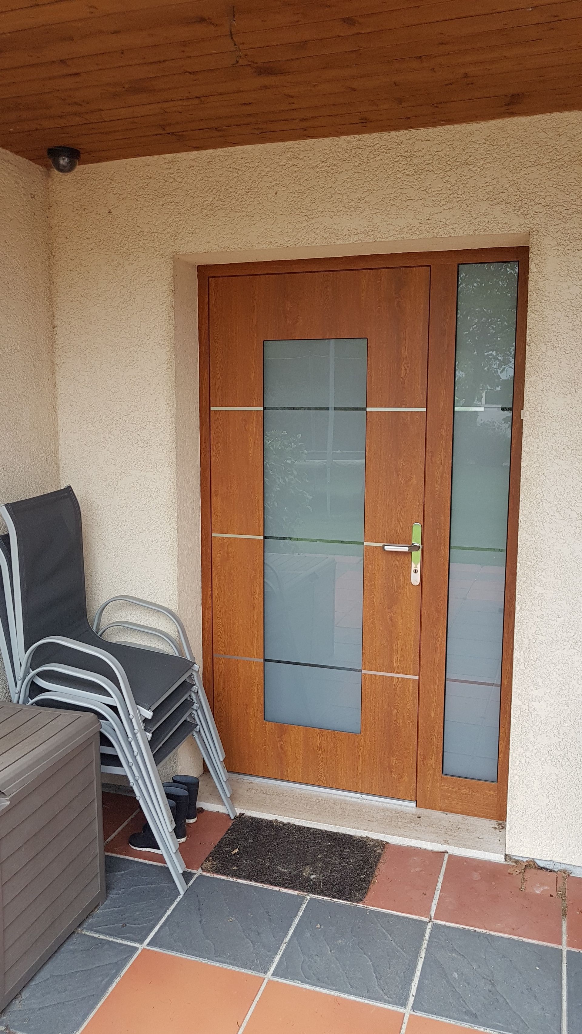 Porte marron avec panneaux de verre dépoli, donnant sur un porche avec des chaises empilées et un paillasson.