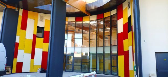 Façade colorée du bâtiment avec une grande entrée vitrée, ornée de panneaux rouges, jaunes et blancs.