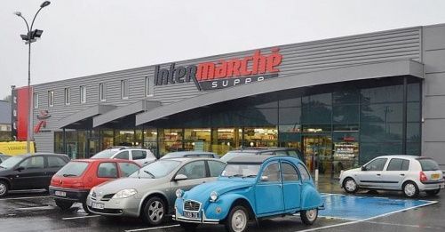 Façade d'un supermarché Intermarché avec des voitures garées par temps de pluie. Une Citroën bleue vintage se distingue.