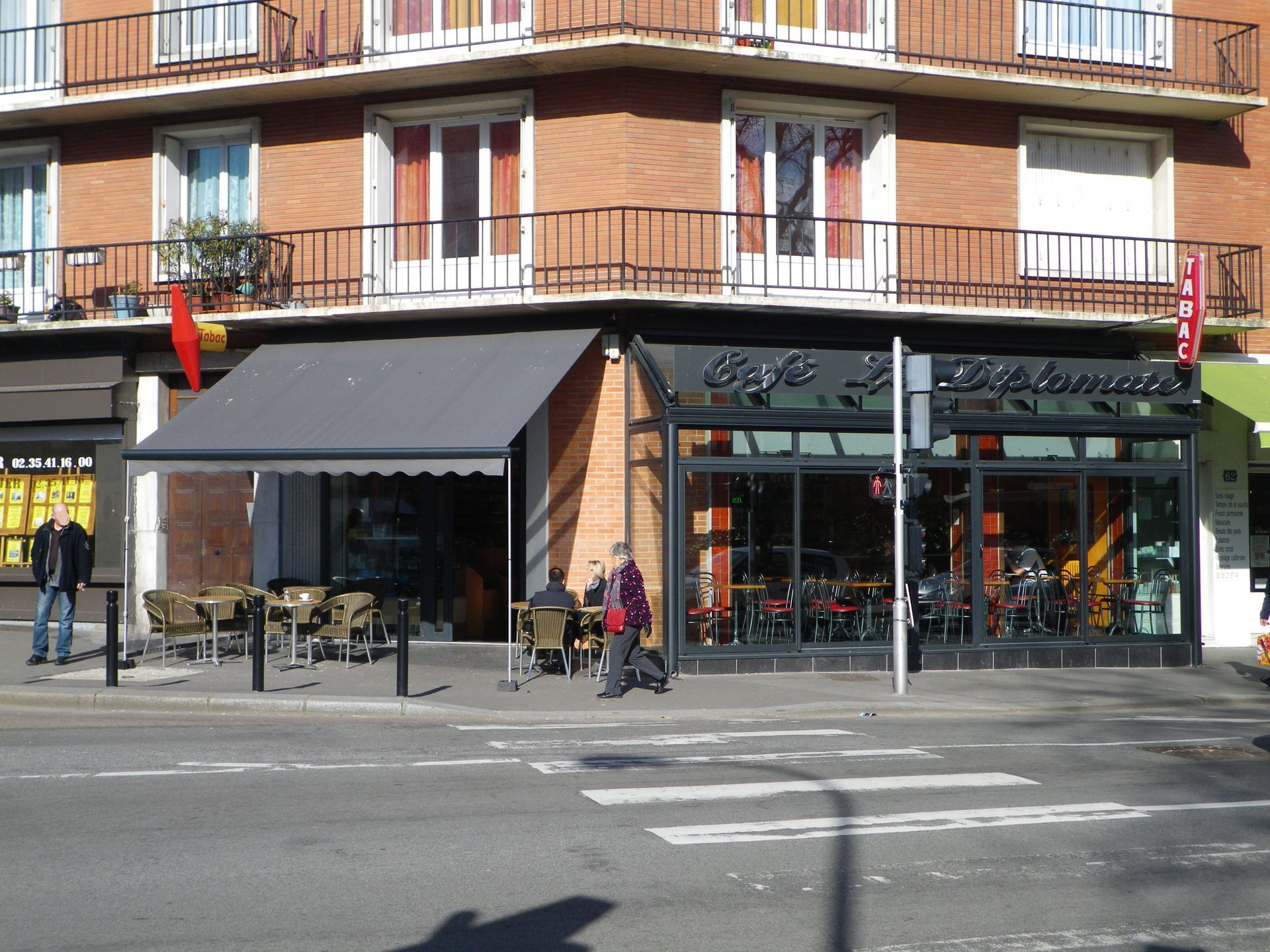 Café d'angle, avec terrasse ombragée par un auvent gris. Immeubles au-dessus, passants à proximité.