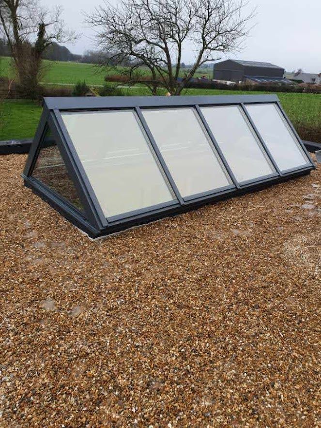 Puits de lumière incliné sur toit de gravier, cadre gris, quatre panneaux de verre, cadre rural avec champs.