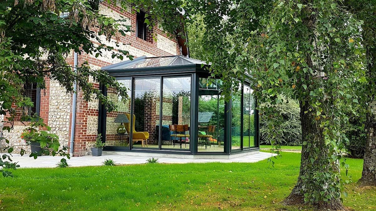 Véranda vitrée attenante à un bâtiment en briques, située dans un jardin arboré.