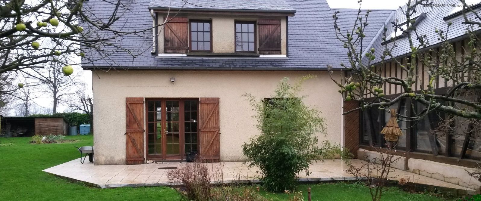 Maison beige avec volets marron, toit en ardoise et pelouse verte.