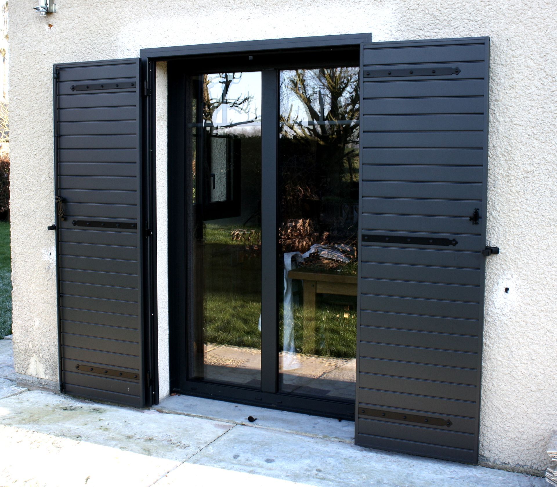 Des volets noirs s'ouvrent de part et d'autre d'une porte vitrée, laissant entrevoir l'extérieur ; la façade du bâtiment est en stuc.