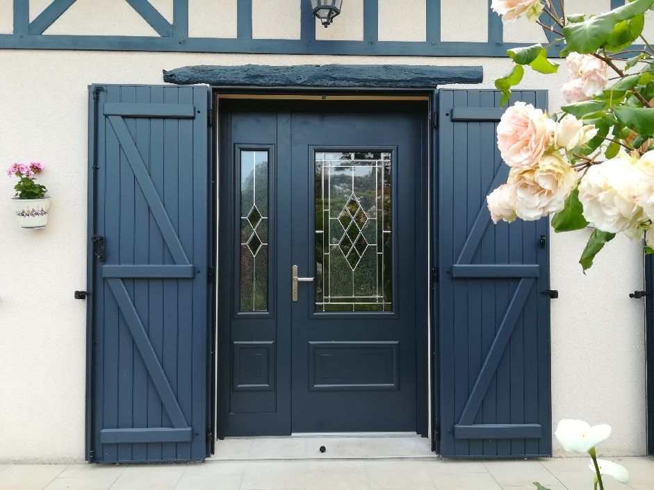 Double porte bleue avec panneaux de verre et volets assortis ; extérieur clair avec un rosier.