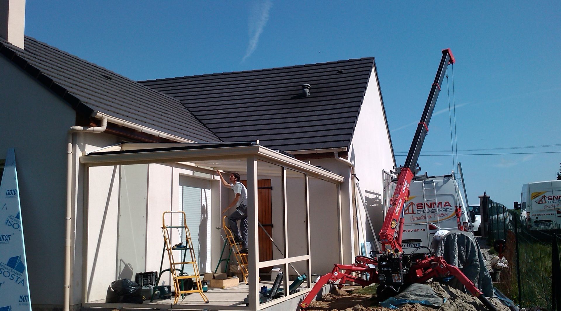 Construction d'une véranda extérieure ; un ouvrier se tient à l'intérieur, une grue est visible.