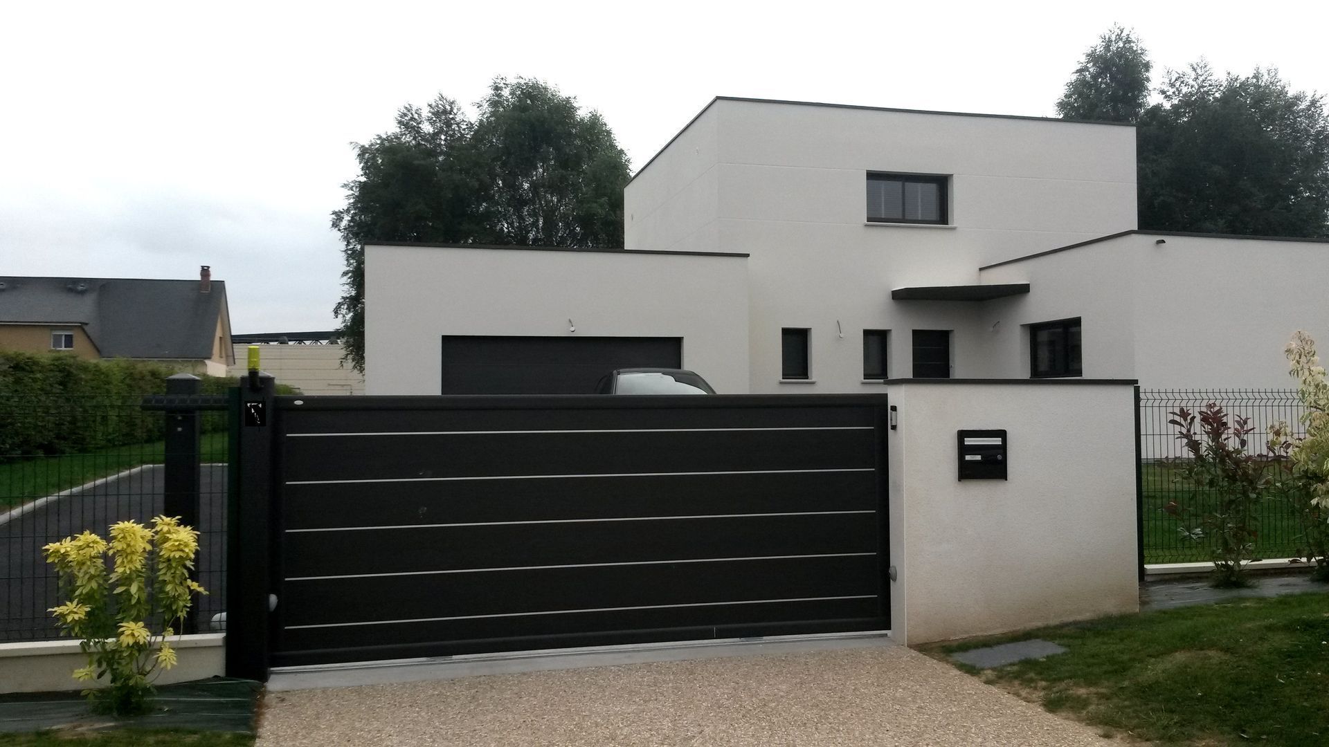 Maison blanche moderne derrière un portail noir ; une voiture est garée dans le garage.