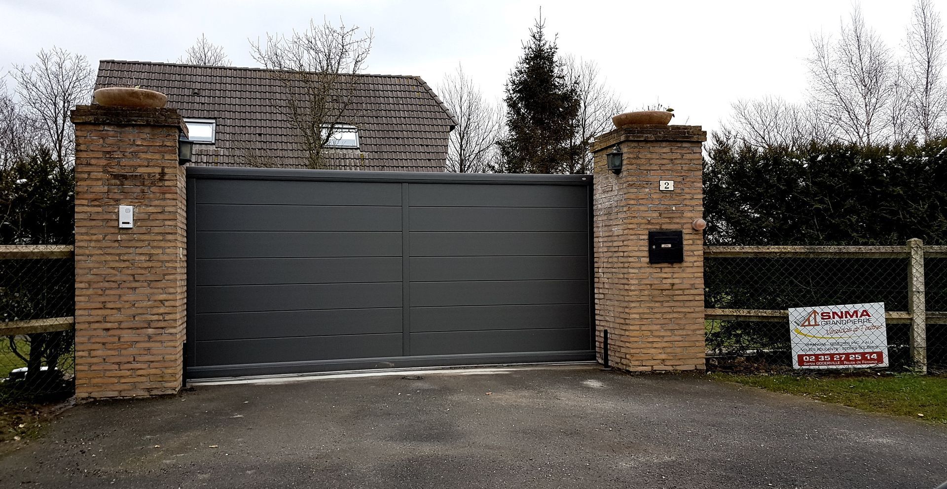 Portail gris entre piliers de briques, donnant accès à une maison. Clôture en bois de chaque côté.