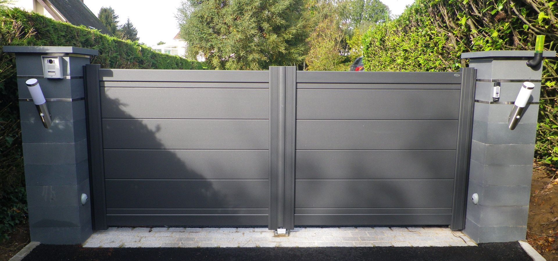 Un portail gris fermé à barreaux horizontaux, flanqué de piliers en béton illuminés. Feuillage vert en arrière-plan.