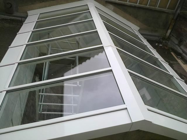 Cage d'ascenseur en verre blanc dans un immeuble avec un escalier intérieur visible.