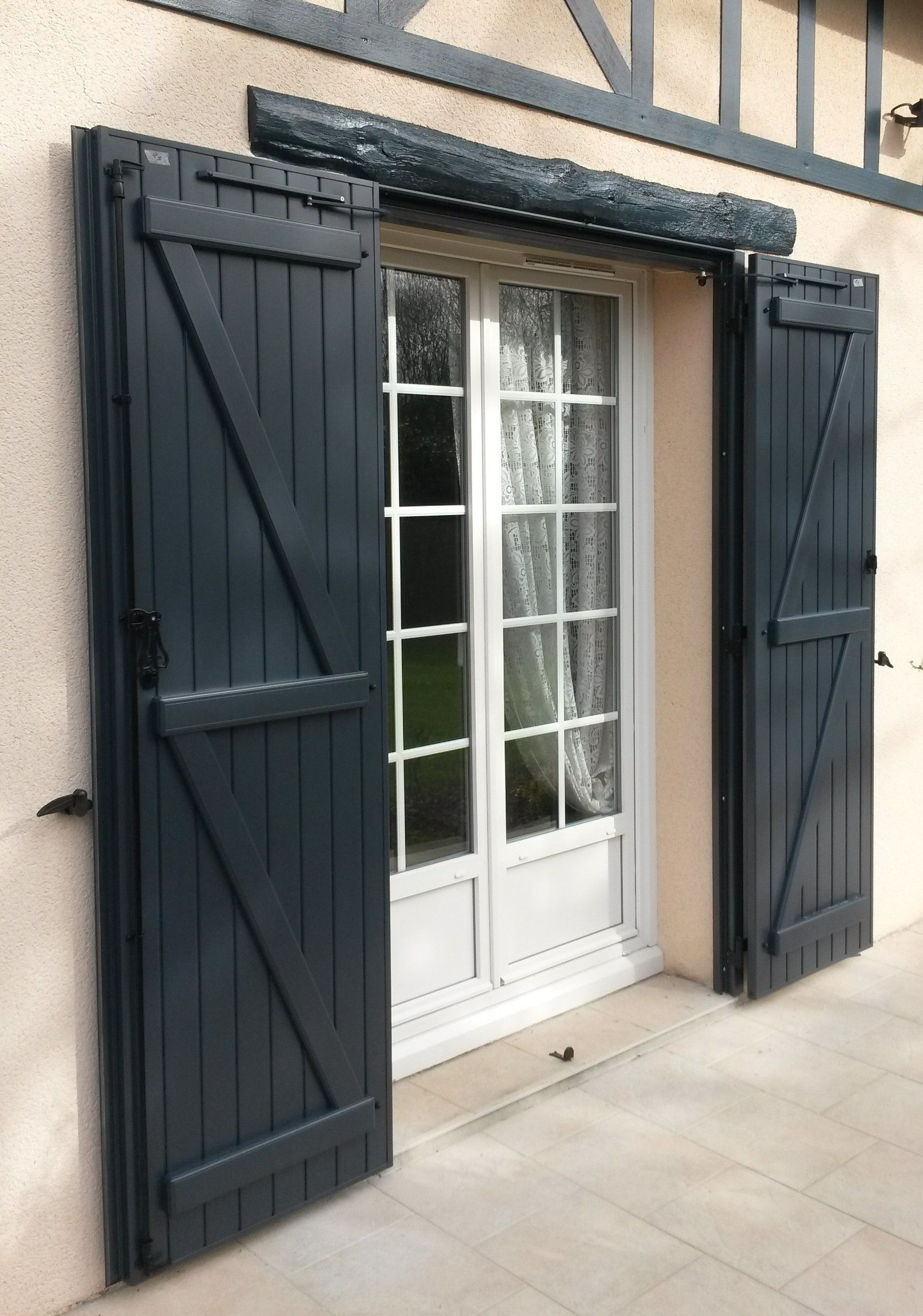 Vue extérieure d'une porte-fenêtre à cadre blanc avec des volets bleu foncé ouverts sur un bâtiment de couleur claire.