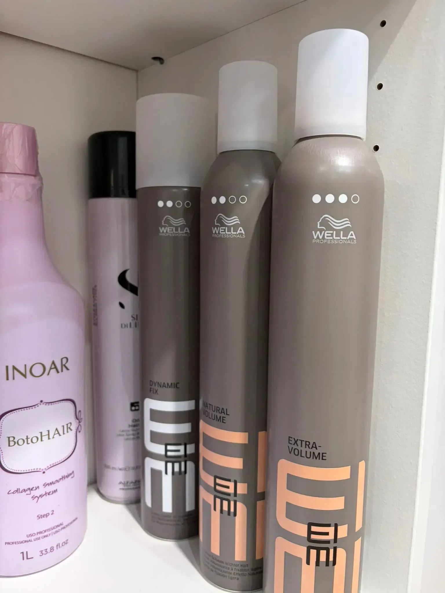 Productos para el cabello en estanterías: botella rosa, lata negra, tres envases de mousse marrón Wella con tapas blancas.