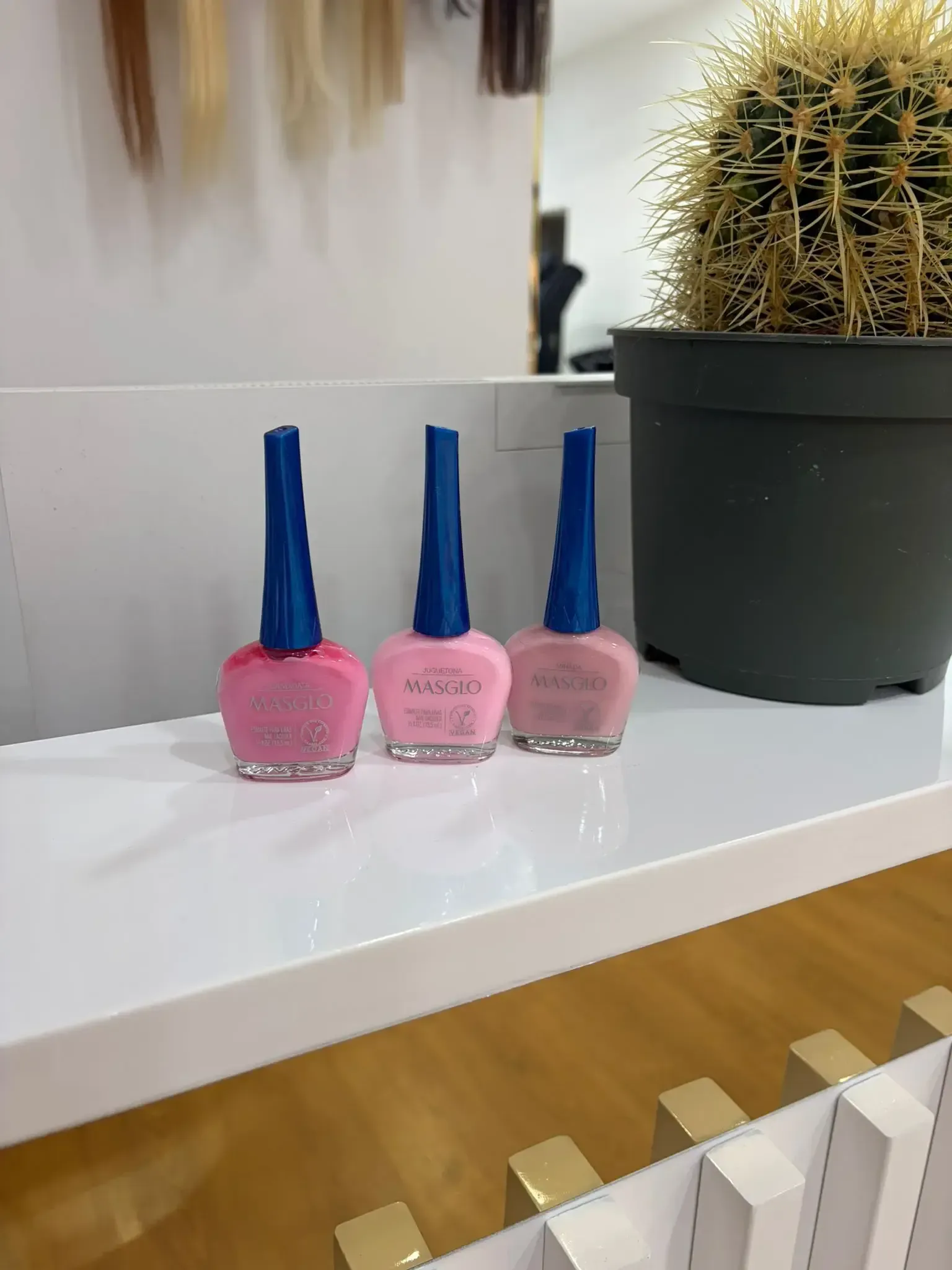 Tres botellas de esmalte de uñas de color rosa con tapas azules en un estante blanco al lado de un cactus.