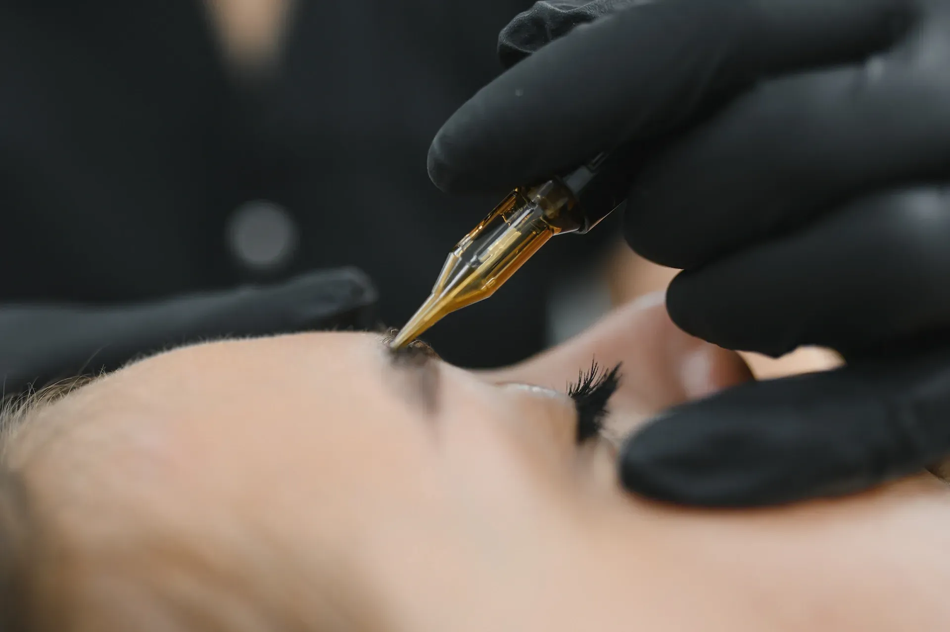 Persona que recibe microblading de cejas; la mano enguantada del artista sostiene la herramienta cerca de la ceja.