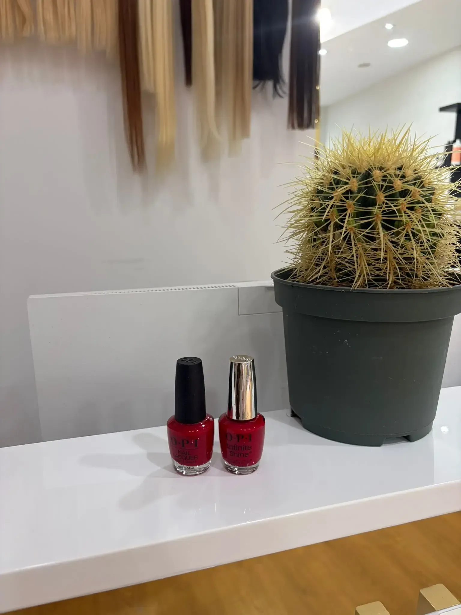 Dos frascos de esmalte de uñas rojo junto a un cactus en una peluquería. Extensiones de cabello colgadas al fondo.