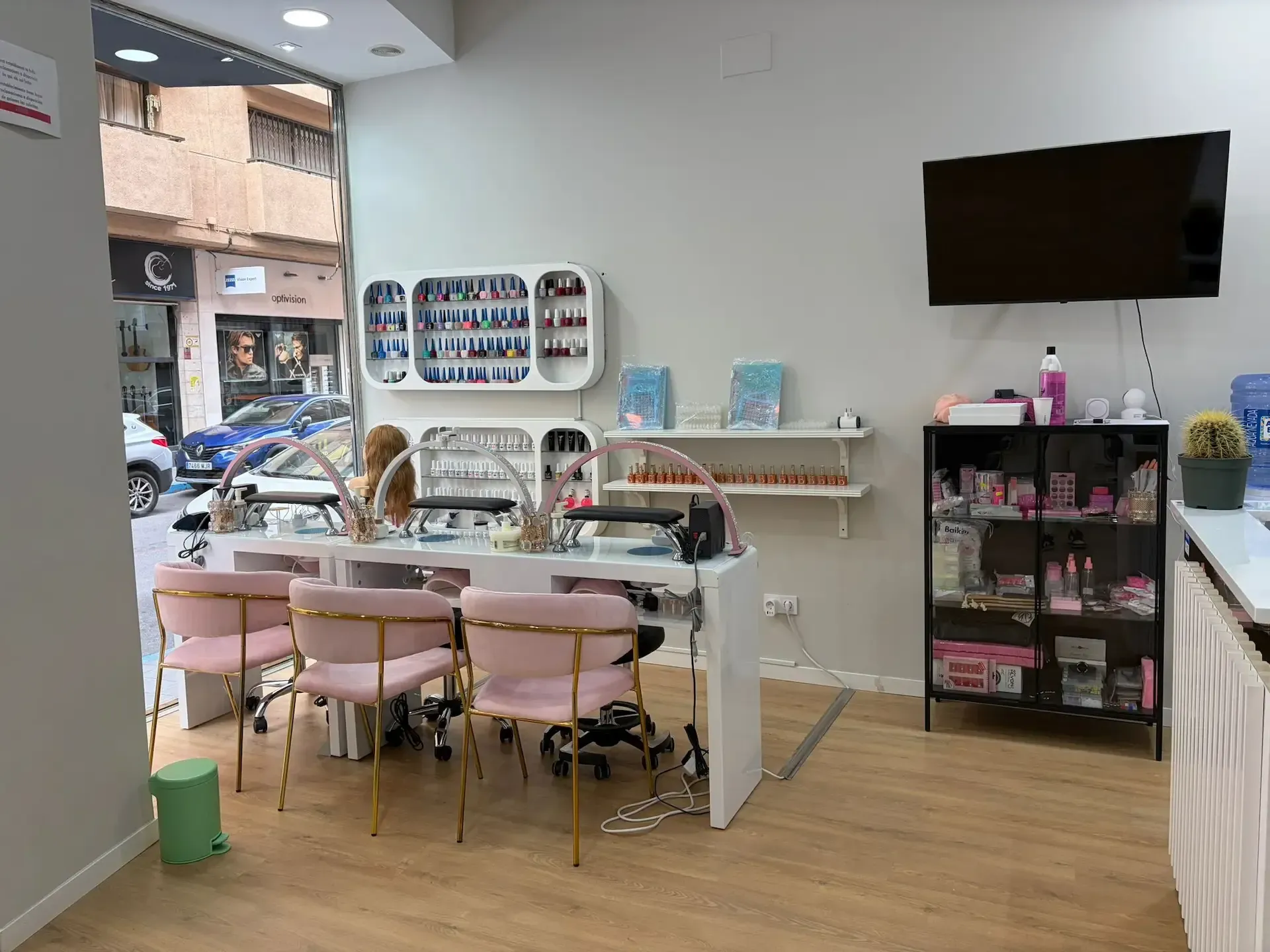 Interior de salón de uñas con mesas para uñas, sillas rosas, exhibiciones de productos y un televisor montado.